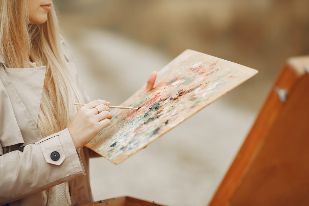 Crop unrecognizable concentrated young female artist in trench coat using paintbrush and mixing colors on palette against wooden easel