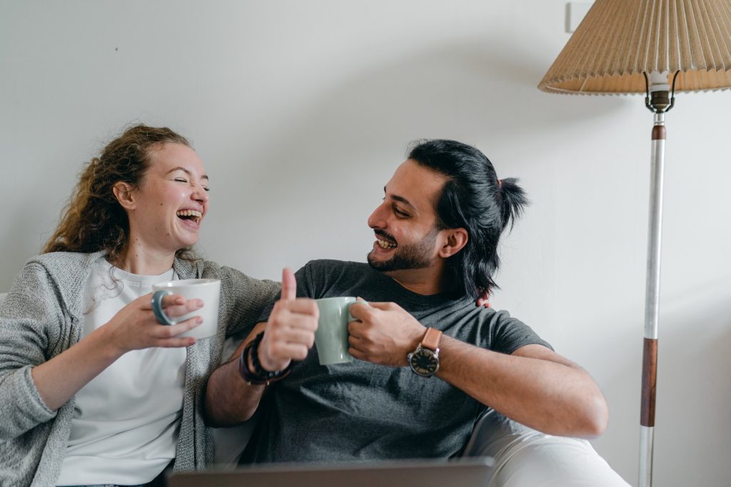 Positive girlfriend in casual wear looking at content ethnic man in smart watch while sitting on cozy sofa with cups of hot beverage and looking at each other