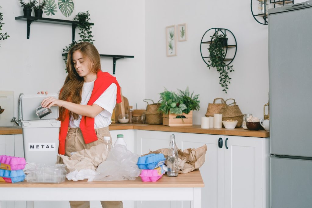 Young responsible female standing in kitchen and putting tin can into bucket while sorting trash