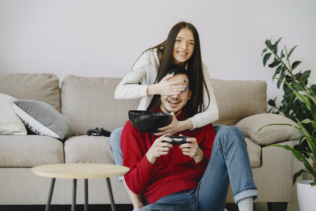 Happy girlfriend covering unrecognizable mans eyes with hand while using game pad and sitting on cozy sofa with bowl at home near potted plant