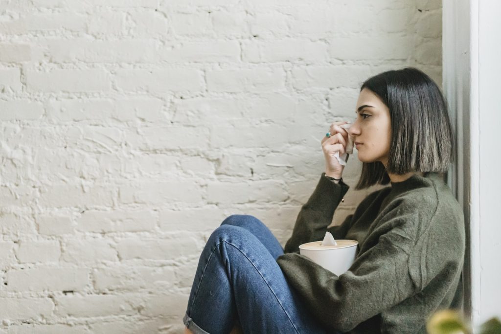 Side view of depressed young ethnic female millennial in warm sweater sitting leaning on wall and wiping tears with tissue at home