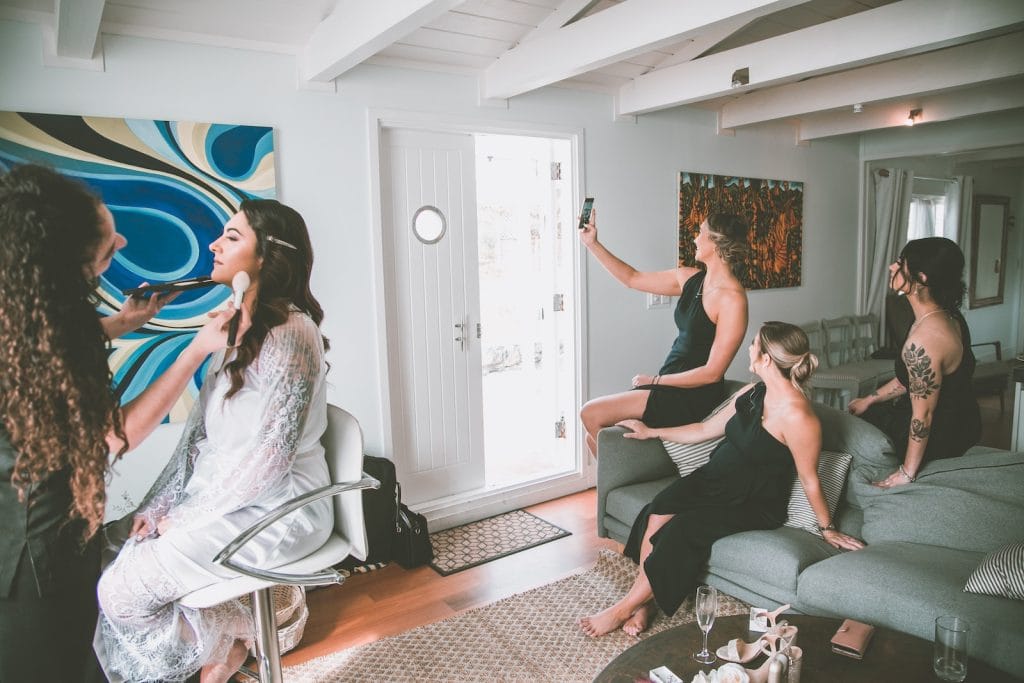 Visagiste doing makeup for charming bride in white robe while bridesmaids in dresses taking selfie on sofa