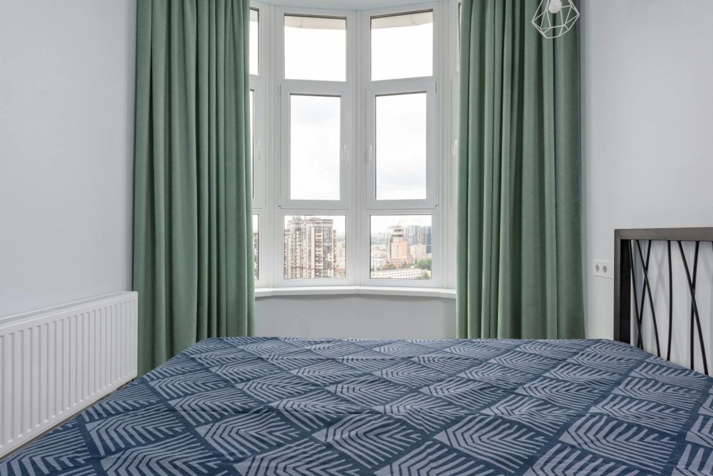 Interior of cozy light bedroom with blue blanket on bed placed against window with green curtain and white walls at home